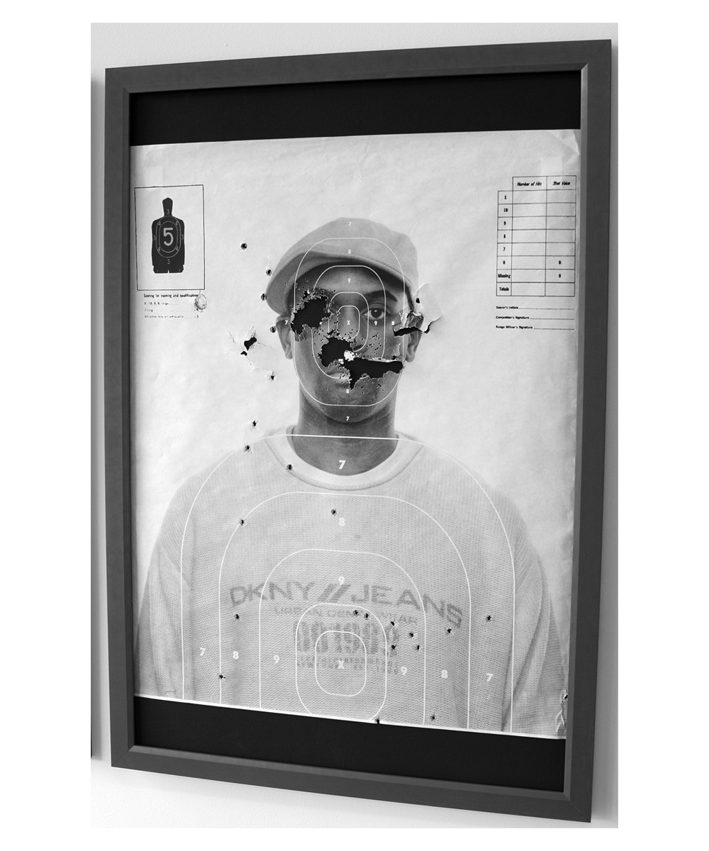 A similar Black-and-white photo of a twenty-something Black man, wearing a DKNY hoodie and Kangol hat, looking directly at the camera, target practice symbols superimposed over his face and chest. He is looking directly at the camera with a neutral expression on his face. Unlike in the first picture, there are multiple bullet holes in the paper around his head and chest; his left eye, nose, and half of his mouth are almost completely shot off. A thin frame holds the bullet-torn piece of paper.