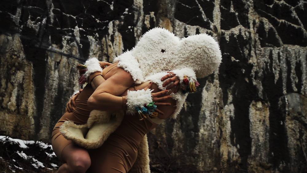 Against a stone wall, two dancers wearing wool masks and beige leotards are locked in fierce embrace once again. This time, one dancer has leapt into the other’s arms, their legs nearly straddling the other body.