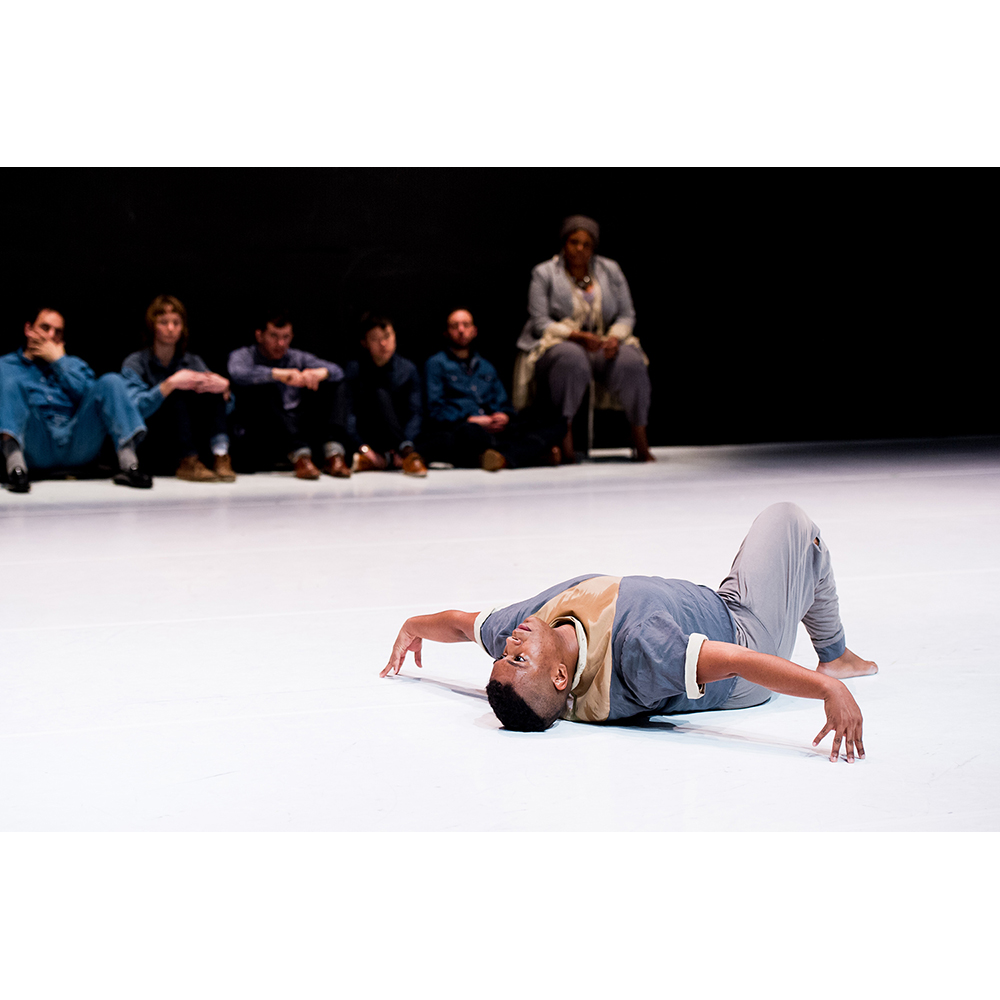 On a white floor, a Black man lays on his back with on leg up and foot planted on the floor. His arms are stretched out in a T. His back is arched. He wears a dark gray t-shirt with white cuffs rolled up, and a shiny section in the middle of his chest in the shape of a V. The V is made of a brown shiny fabric. He wears gray pants. In the background, a blurry row of four people wearing shades of blue denim, sit on the ground and watch. There is also a woman, wearing gray, sitting on a chair and watching. Everyone looks on seriously. 