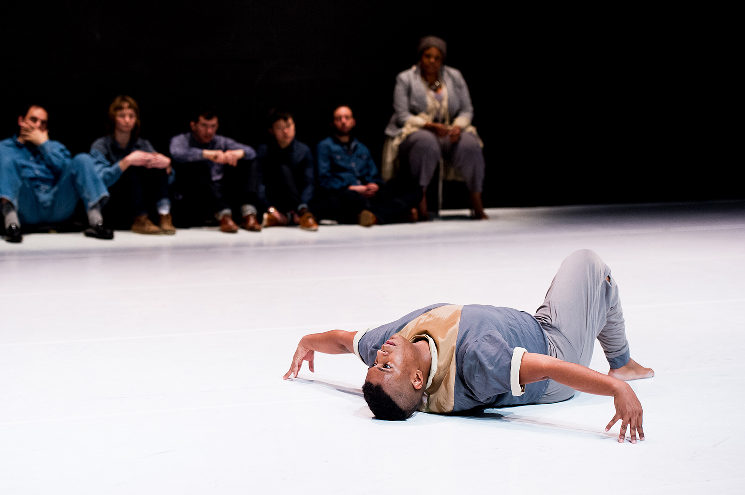 On a white floor, a Black man lays on his back with on leg up and foot planted on the floor. His arms are stretched out in a T. His back is arched. He wears a dark gray t-shirt with white cuffs rolled up, and a shiny section in the middle of his chest in the shape of a V. The V is made of a brown shiny fabric. He wears gray pants. In the background, a blurry row of four people wearing shades of blue denim, sit on the ground and watch. There is also a woman, wearing gray, sitting on a chair and watching. Everyone looks on seriously.